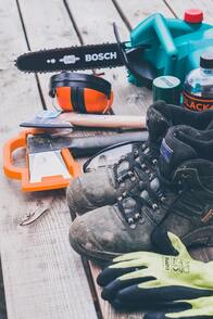 A picture of green gloves, brown work boots, orange tools, and a dark green chainsaw laid out on a porch.